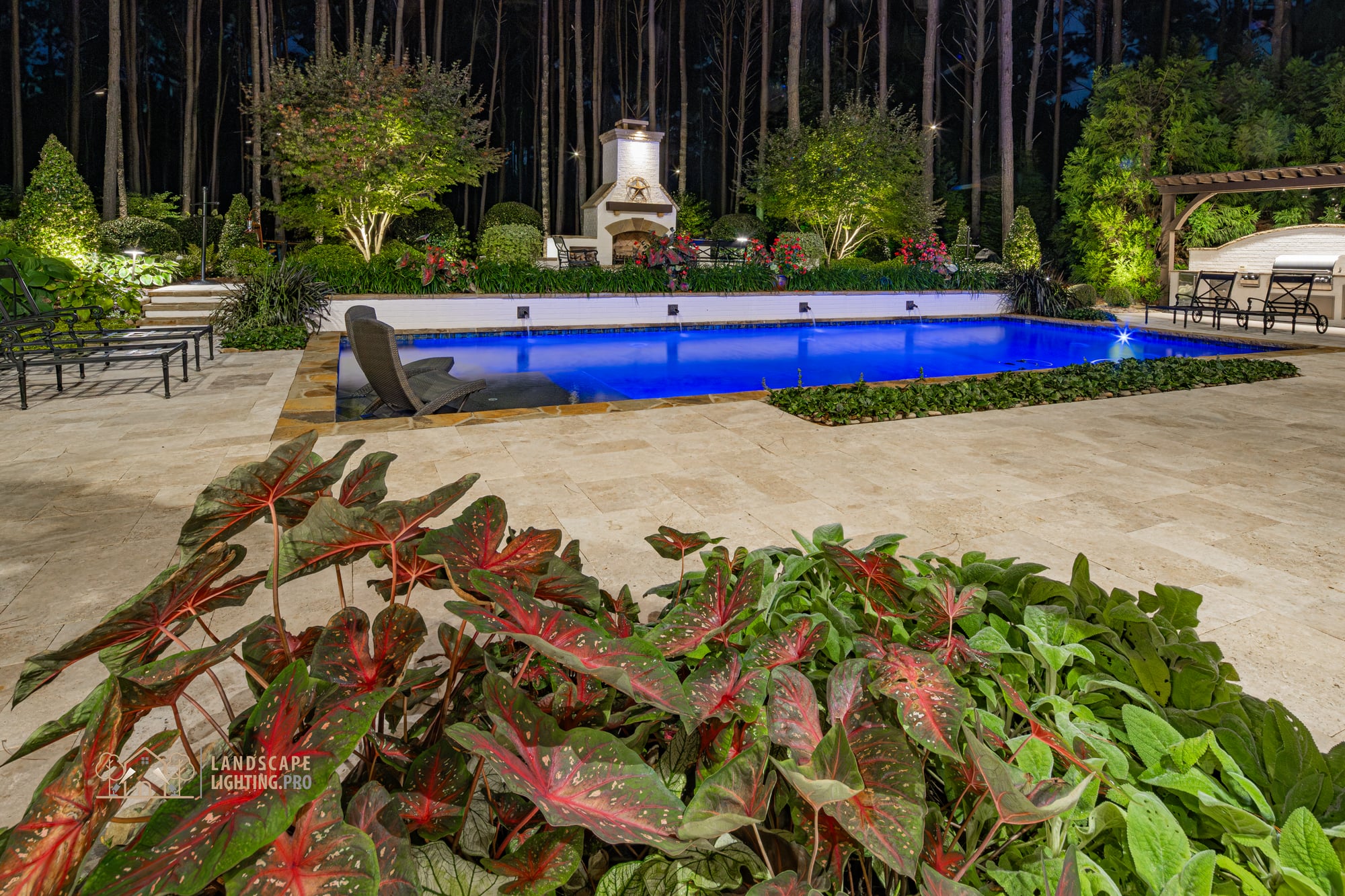 A rectangular pool at dusk with an outdoor fireplace, dining terrace, and tropical plantings, all illuminated as a single composition